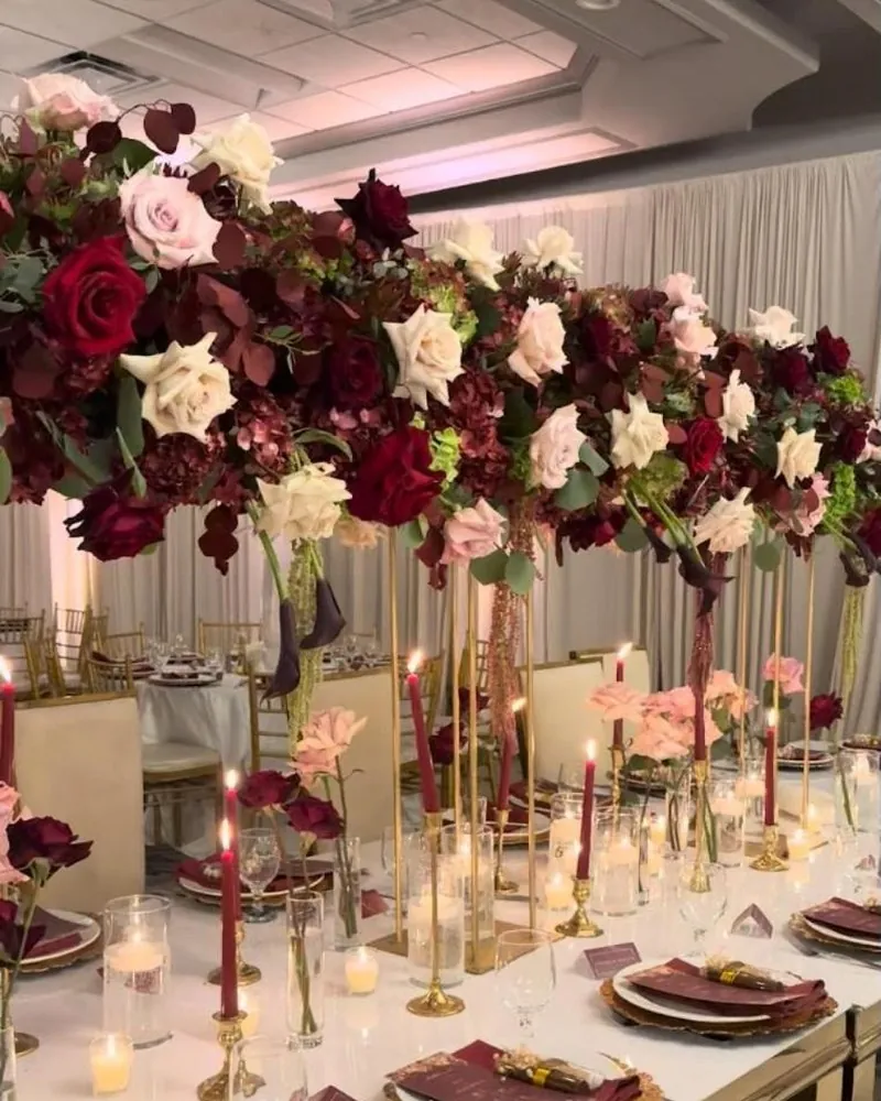 Long table lined with tall burgundy and blush rose arrangements, taper candles and gold chargers