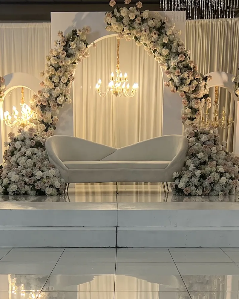 White stage with a floral arch of ivory and blush roses, a curved loveseat and a crystal chandelier